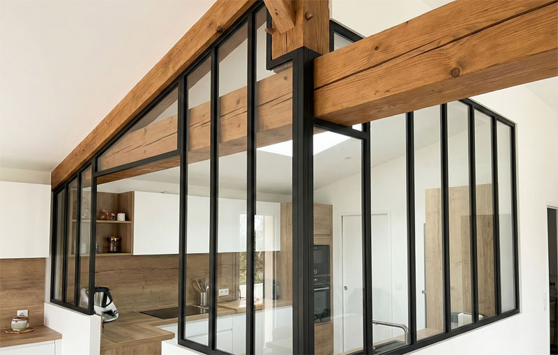 Verrière de cuisine en acier noir sur mesure, contournant des poutres en bois massives et posée sur un muret blanc pour délimiter l'espace.