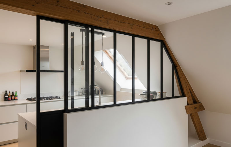 Verrière de cuisine sur mesure en acier noir avec profilés fins, parfaitement intégrée sous un plafond rampant avec poutre apparente.