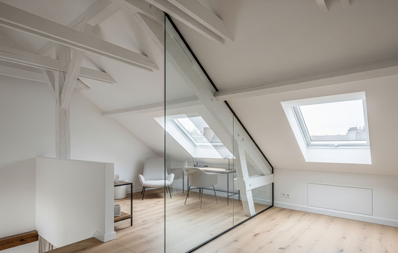 Une verrière sous pente sur mesure avec un cadre en métal noir installée dans une mezzanine mansardée lumineuse. La cloison vitrée suit parfaitement l'inclinaison du toit, séparant les espaces tout en laissant passer la lumière naturelle dans des combles aménagés au style moderne.
