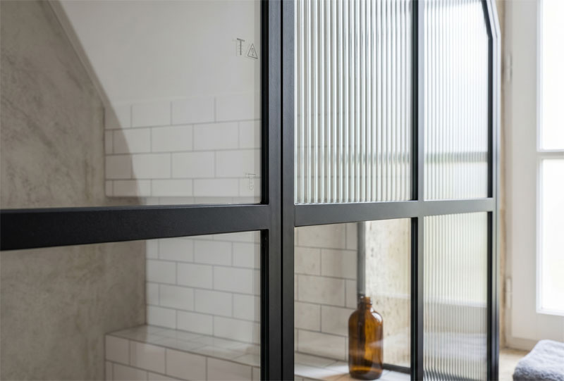 Détail des finitions d'une paroi de douche montrant l'association d'un vitrage flûté (strié) et d'un vitrage clair dans un châssis en acier thermolaqué.