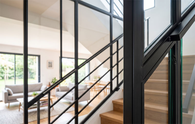 Verrière d'escalier sur mesure en acier noir mat, séparant une montée d'escalier en bois d'un salon moderne et lumineux.
